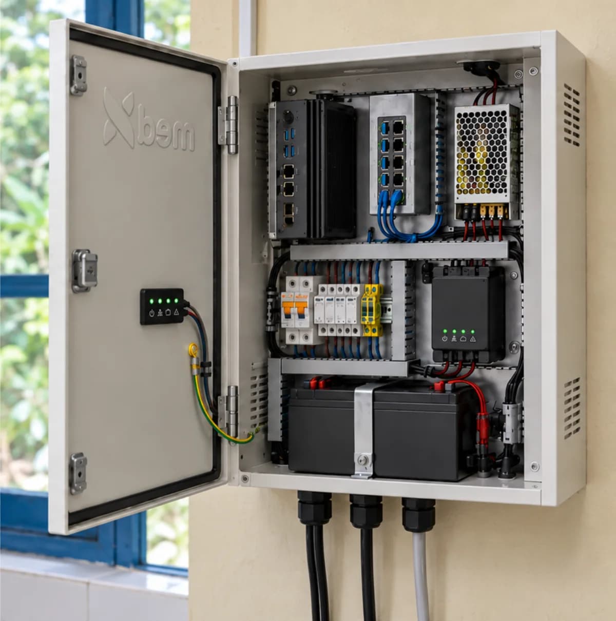 The cabinet open on a laboratory wall, showing the mini PC and switch on the top shelf, protection devices on the DIN rail below, and the batteries at the bottom